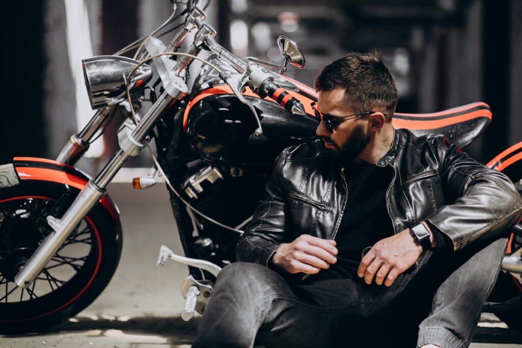 A handsome man with a beard and sunglasses in a leather jacket sitting next to a custom motorcycle.